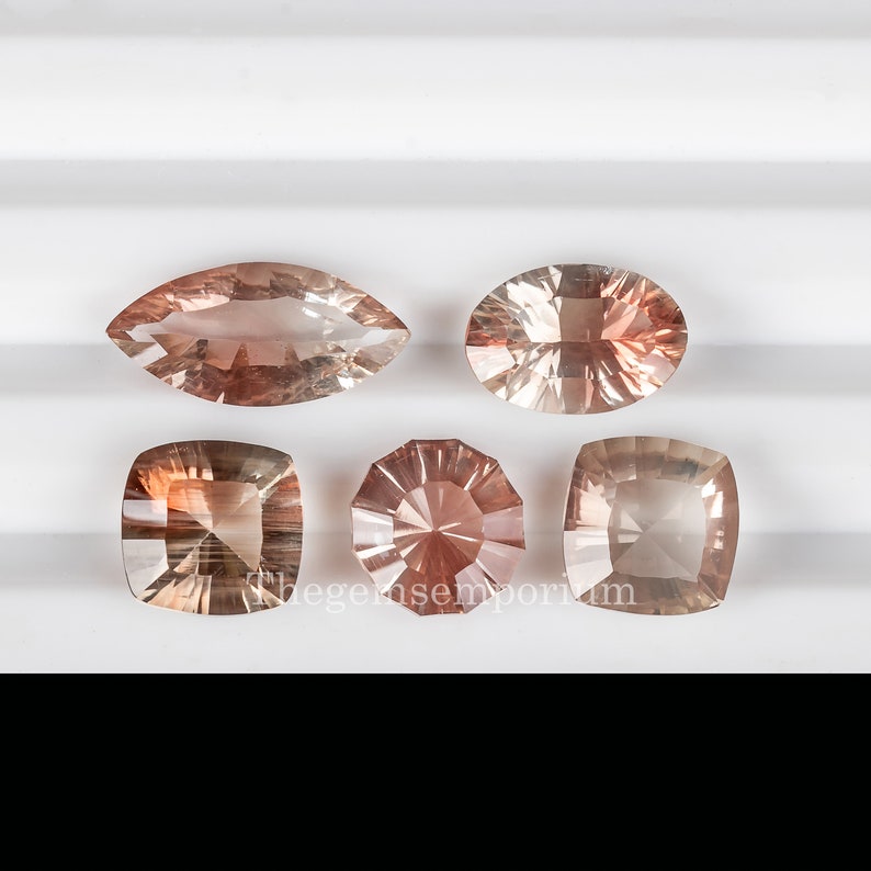 May include: Five sunstone gemstones in various shapes and sizes. The gemstones are a light orange color with a slight pink hue. The gemstones are displayed on a white surface.