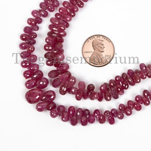 May include: A strand of deep red ruby gemstone beads, each bead is faceted and shaped like a teardrop. The beads are strung on a clear thread and are approximately the size of a US penny.
