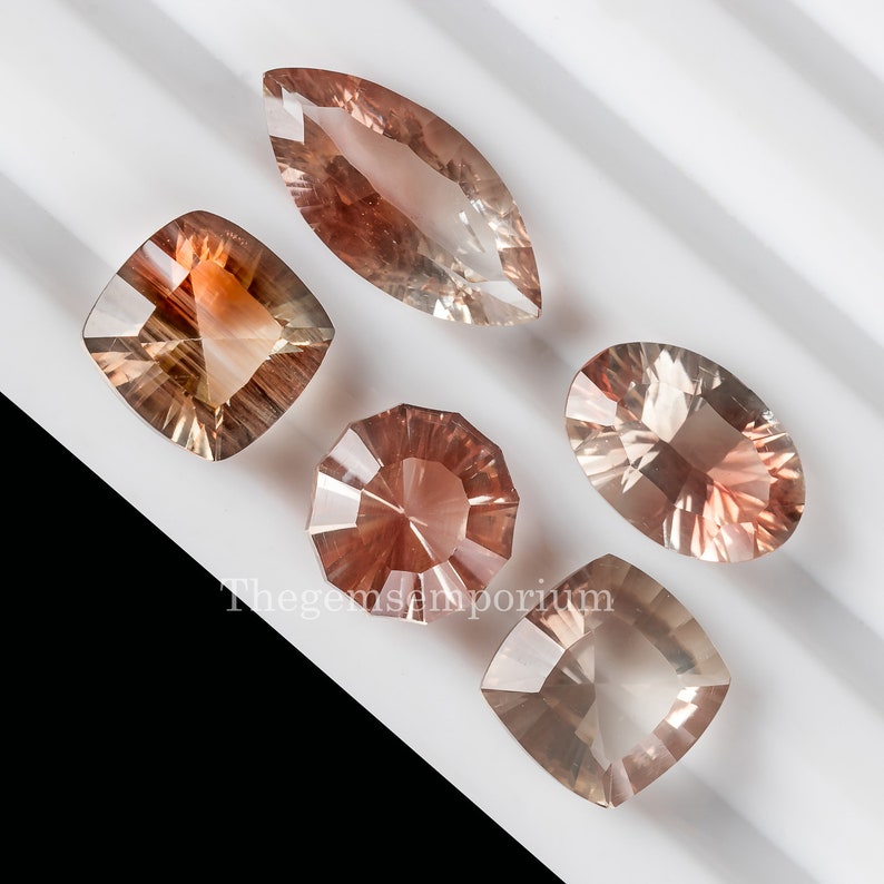 May include: Five sunstone gemstones in various shapes and sizes. The gemstones are a mix of orange, red, and clear colors. The gemstones are displayed on a white surface.