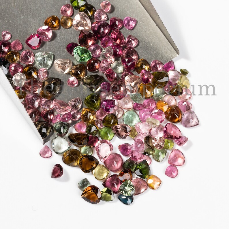 May include: A large collection of loose, faceted, pear-shaped gemstones in various shades of pink, green, brown, and clear. The gemstones are scattered on a silver tray and a white surface.