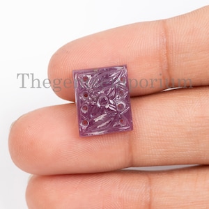 May include: A carved pink gemstone with a floral design. The gemstone is square shaped and has holes drilled around the edges.