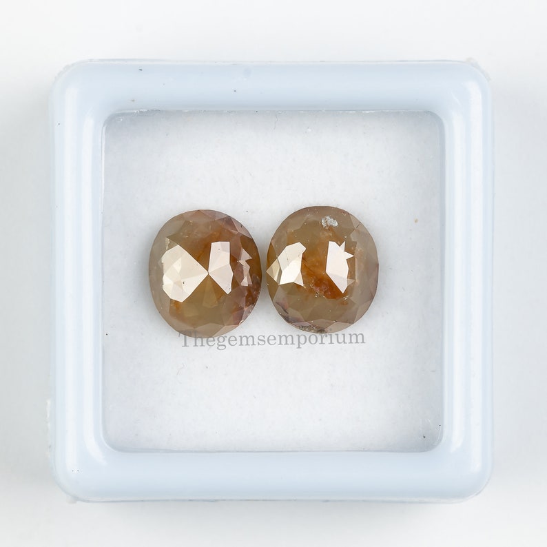 May include: Two brown, round, faceted diamonds in a white box. Thegemsemporium is printed on the bottom of the box.