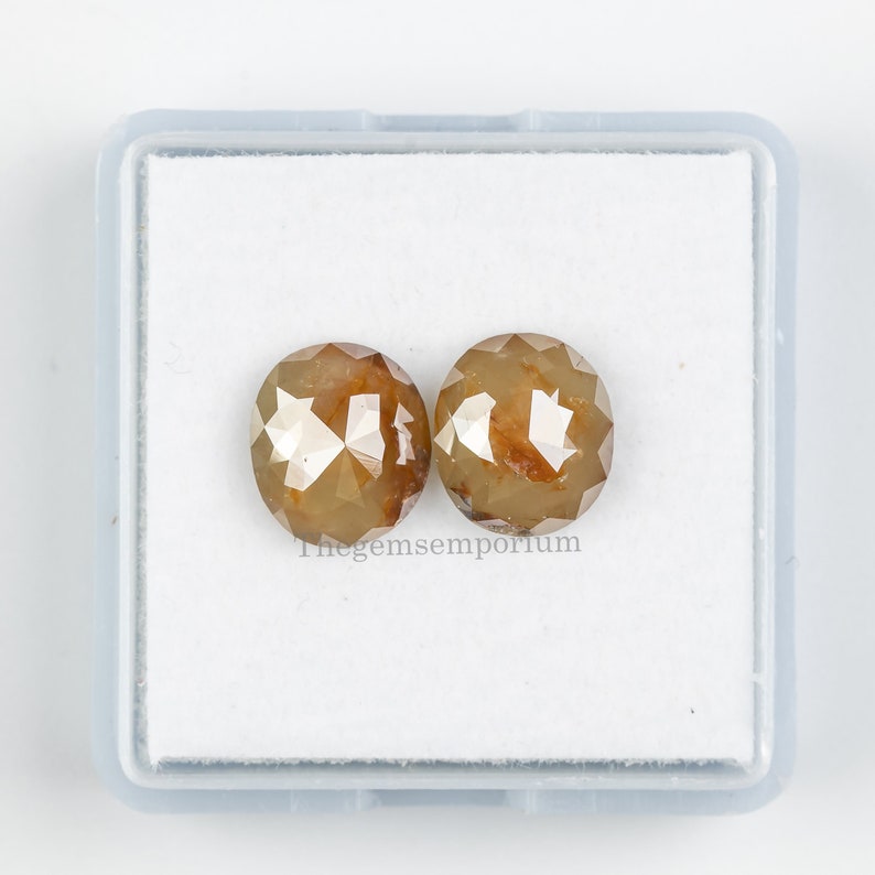 May include: Two round, faceted, brown diamonds in a white box. Thegemsemporium is printed on the bottom of the box.
