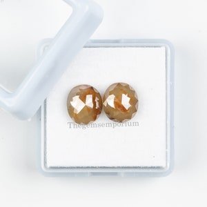 May include: Two brown round diamonds in a white box. The diamonds have a faceted cut and are set in a white box. Thegemsemporium is printed on the box.