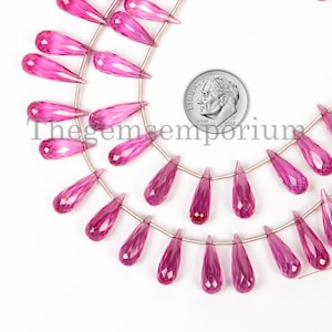 Pode incluir: Um fio de contas de pedra preciosa rosa facetadas em forma de lágrima, enfiadas em um fio de prata. As contas medem aproximadamente 2,5 cm de comprimento e são mostradas ao lado de uma moeda de dez cêntimos de euro para a escala.