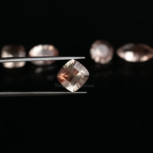May include: A square-shaped, light brown gemstone with a shimmery, iridescent finish. The gemstone is held in tweezers against a black background.