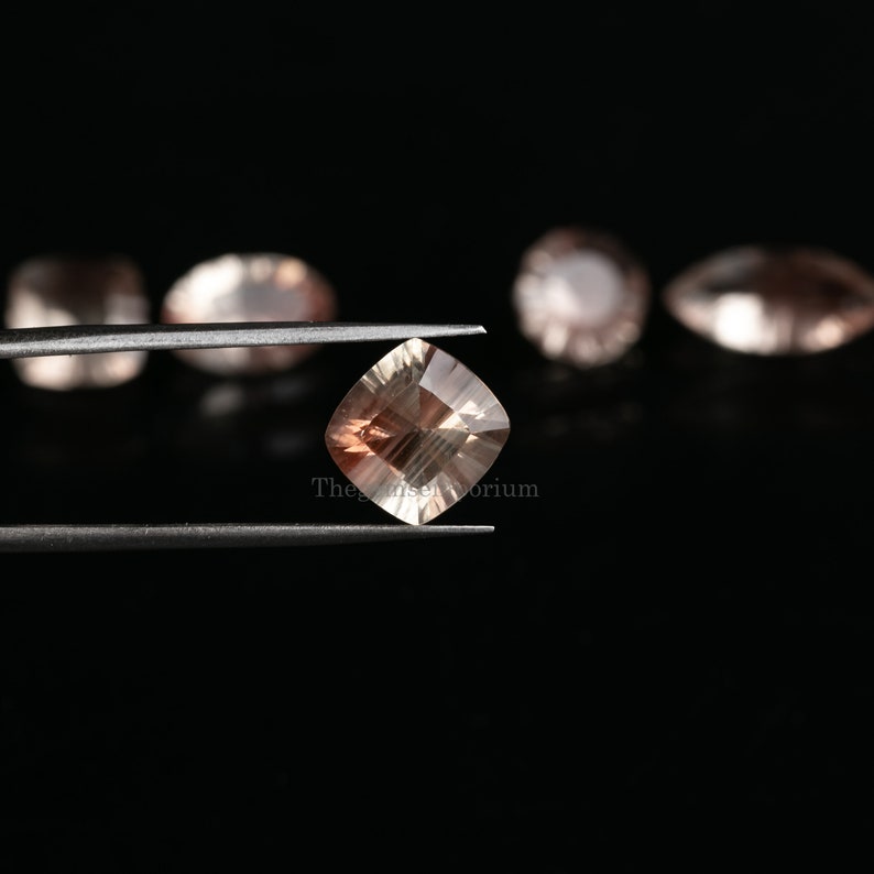 May include: A square-shaped, light brown gemstone with a shimmery, iridescent finish. The gemstone is held in tweezers against a black background.