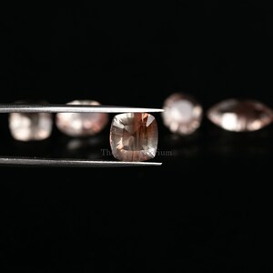 May include: A close-up of a sunstone gemstone held in tweezers. The gemstone is a light brown color with a square shape and a faceted cut. The gemstone is surrounded by other sunstones in the background.