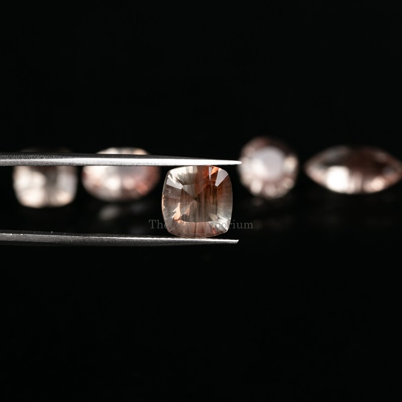 May include: A close-up of a sunstone gemstone held in tweezers. The gemstone is a light brown color with a square shape and a faceted cut. The gemstone is surrounded by other sunstones in the background.