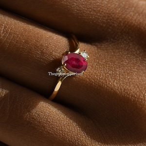 May include: A gold ring with a red oval ruby gemstone and two smaller white diamond accents. The ring has a curved band.