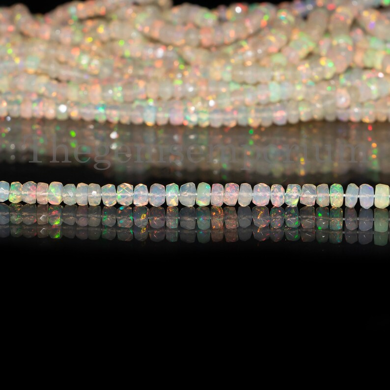 May include: A strand of white opal beads with iridescent flashes of color. The beads are faceted and have a rectangular shape. The beads are strung on a clear thread and are lying on a black surface.