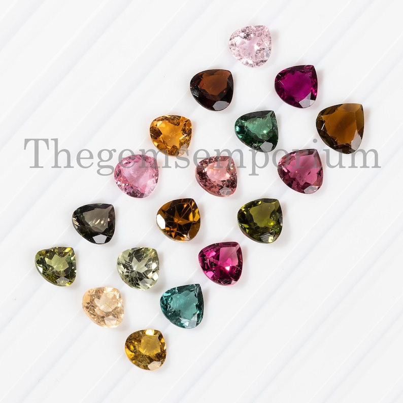 May include: Assortment of 16 heart-shaped gemstones in various colors, including pink, green, brown, and yellow. The gemstones are arranged in a diagonal line on a white surface. Thegemsemporium is printed in black text on the surface.