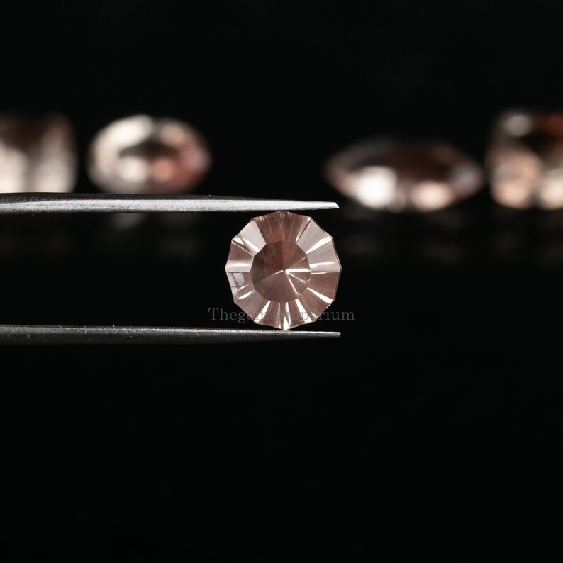 May include: A light pink gemstone with a faceted, starburst design is held in tweezers against a black background.