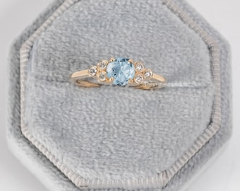 May include: A delicate gold ring with a light blue gemstone surrounded by smaller clear gemstones. The ring is displayed in a gray velvet ring box.