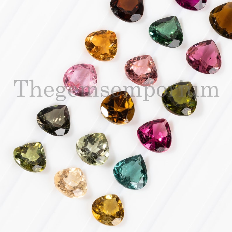 May include: Assortment of heart-shaped gemstones in various colors, including pink, green, brown, and yellow. The gemstones are arranged on a white surface.