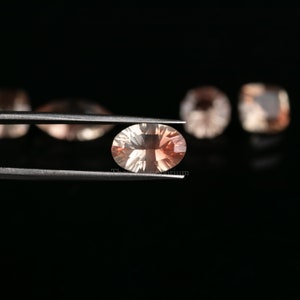 May include: A light orange oval-shaped gemstone held by tweezers against a black background. The gemstone has a slight shimmer and is surrounded by other out-of-focus gemstones.
