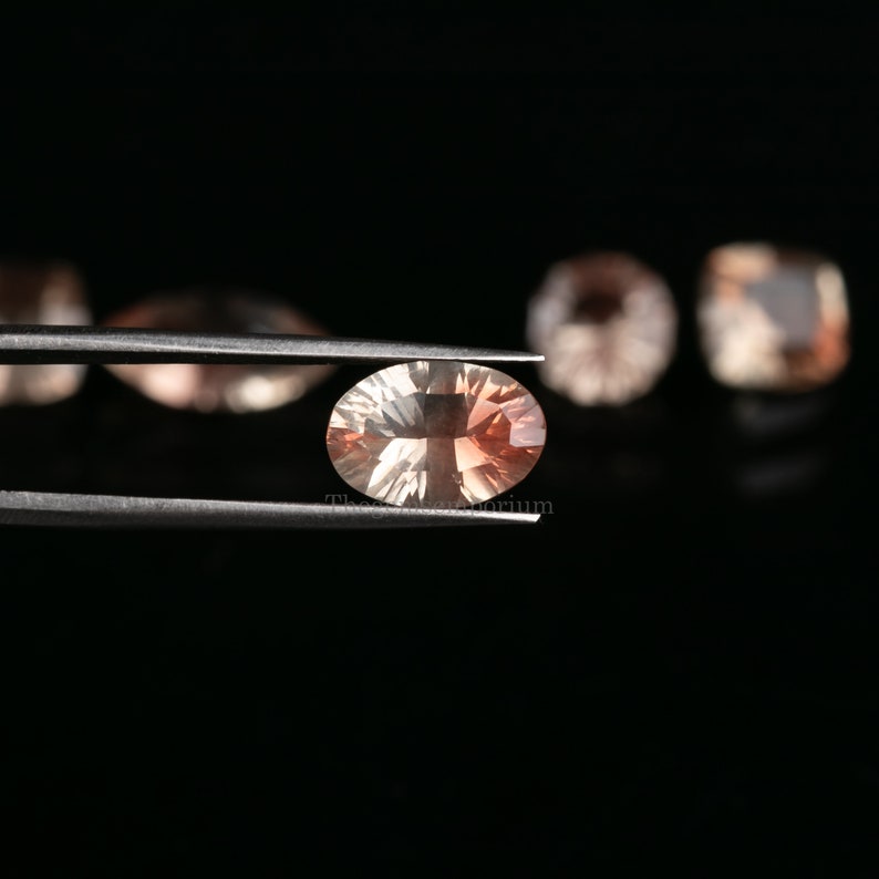 May include: A light orange oval-shaped gemstone held by tweezers against a black background. The gemstone has a slight shimmer and is surrounded by other out-of-focus gemstones.