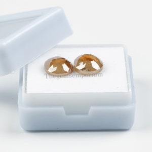 May include: Two brown, round, faceted diamonds in a white box. Thegemsemporium is printed on the box.