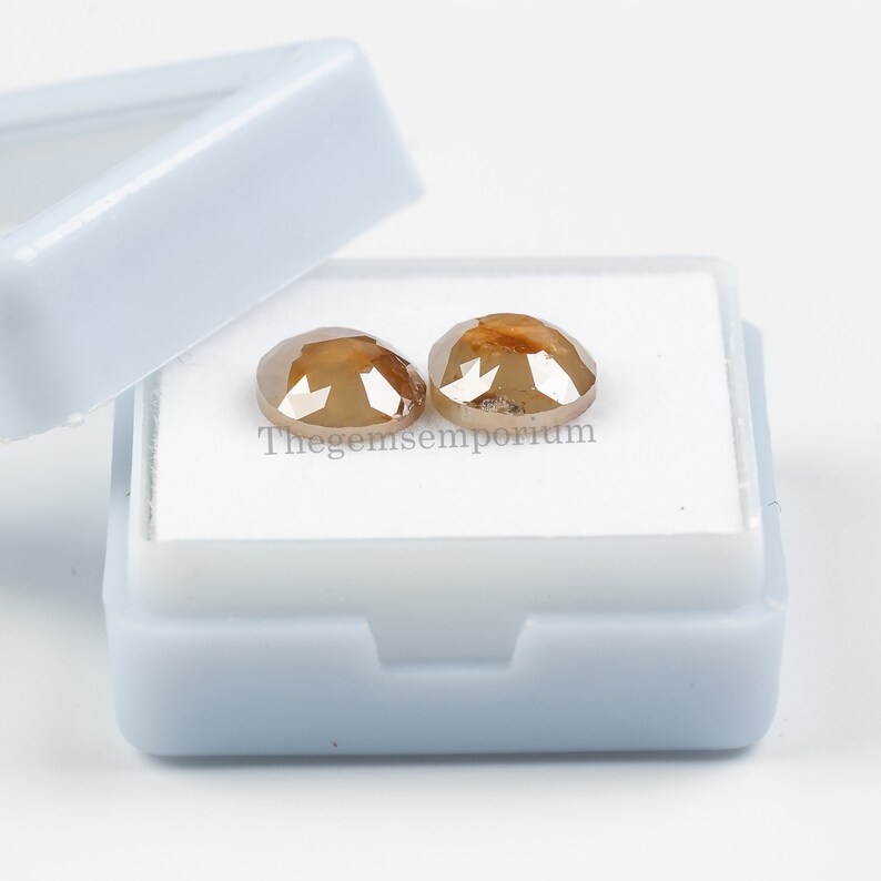 May include: Two brown, round, faceted diamonds in a white box. Thegemsemporium is printed on the box.