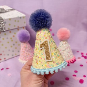 May include: A cone-shaped party hat with a yellow glittery surface covered in multicolored confetti dots. The hat features a dark blue pom-pom on top, a light blue pom-pom trim at the base, and a gold glittery number one centered on the front.