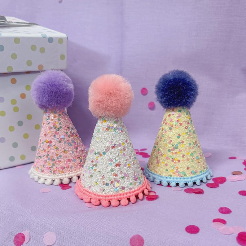 May include: Three cone-shaped party hats featuring glittery surfaces with multicolored confetti accents. Each hat is topped with a large, fluffy pom-pom in purple, pink, or dark blue, and finished with a small pom-pom trim along the bottom edge.