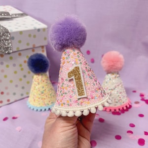 May include: A pink glittery cone-shaped party hat featuring a gold glittery number one, a large purple pom-pom on top, and a white pom-pom trim along the bottom edge. Two additional smaller party hats with pom-poms are visible in the background.