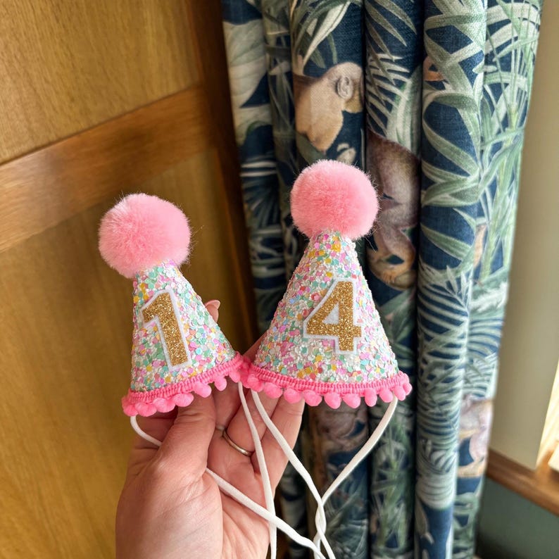 May include: Two cone-shaped party hats covered in multicolored sequins with a pink pom-pom on top and a pink pom-pom trim at the base. One hat features a gold glitter number 1, and the other hat features a gold glitter number 4, each with white elastic straps.