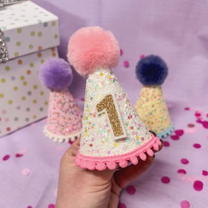 May include: A cone-shaped party hat covered in white glitter with a large gold glitter number one attached to the front. The hat features a soft pink pom-pom on the top and a pink pom-pom trim along the bottom edge, held in a hand.