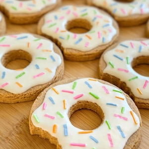 May include: Several round, golden-brown cookies shaped like donuts, each topped with a smooth layer of white icing and scattered with small, colorful sprinkles in shades of pink, blue, green, and orange, arranged in a group on a wooden surface.