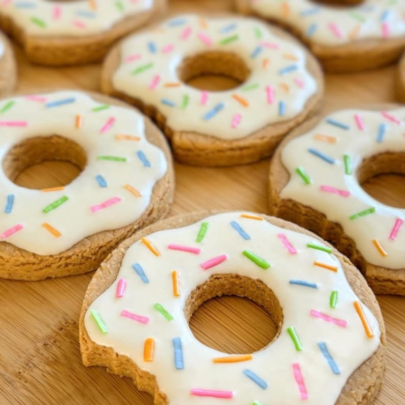 May include: Several round, golden-brown cookies shaped like donuts, each topped with a smooth layer of white icing and scattered with small, colorful sprinkles in shades of pink, blue, green, and orange, arranged in a group on a wooden surface.