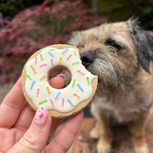 May include: A round, tan-colored dog treat shaped like a donut with a bite taken out of the side. The treat features a smooth white icing layer on top decorated with small, thin, multi-colored sprinkles in shades of pink, blue, green, and orange.