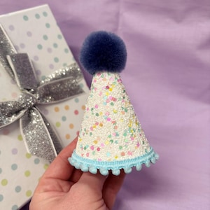 May include: A conical party hat featuring a white textured surface covered in multicolored glitter, topped with a dark blue pom-pom. The base of the hat is trimmed with a light blue pom-pom fringe. The hat is held against a white gift box.