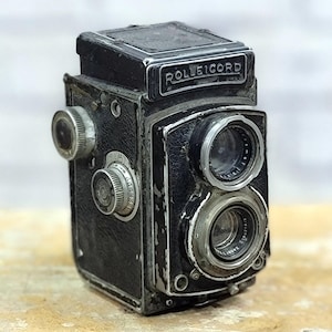 Puede incluir: Cámara Rolleicord negra vintage con detalles plateados. La cámara presenta dos lentes, diales y la palabra "Rolleicord" en la parte superior. Es un diseño clásico de cámara réflex de doble lente, que muestra una estética retro.