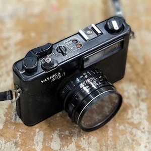 May include: A black Yashica MG-1 35mm film camera with a lens. The camera has a silver lens ring and a black strap. The camera body has the text "Yashica MG-1" printed on the front. The camera is on a wooden surface.
