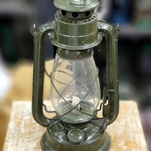 Könnte beinhalten: Eine olivgrüne Vintage-Laterne mit einem durchsichtigen Glaszylinder und einem Metallgriff. Die Laterne hat einen runden Sockel und einen gewölbten Deckel mit Belüftungslöchern. Die Laterne steht auf einer Holzoberfläche.