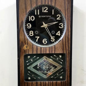 May include: Vintage rectangular wall clock with a wood grain frame. The clock face is black with white numerals and hands, and the word "SCIENTIFIC" is printed above the center. The lower section features a decorative geometric design.