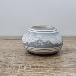 May include: A small, grey and white ceramic pot with a mountain scene design. The pot has a rounded shape and is decorated with white stripes.