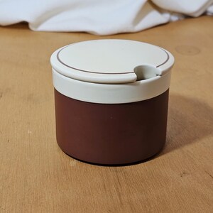 May include: A small, round ceramic container with a lid. The container is a deep brown colour with a white band around the top. The lid is white with a brown trim and a small notch for a spoon. The container is likely for storing sugar or spices.