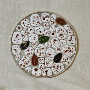 May include: A circular coaster with a silver edge, featuring a mosaic design. The mosaic is composed of white tiles with brown speckles, and leaf-shaped tiles in green, brown, and black. The coaster is approximately 10 cm in diameter.