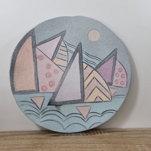 May include: A round ceramic plate with a blue and white background. The plate features a geometric design of pink, peach, and purple triangles with black outlines. The triangles are arranged to resemble a cityscape.
