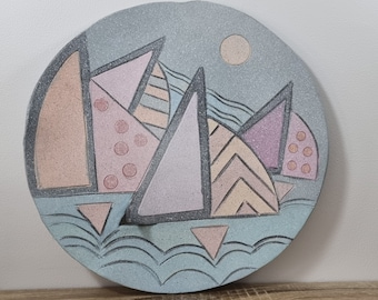 May include: A round ceramic plate with a blue and white background. The plate features a geometric design of pink, peach, and purple triangles with black outlines. The triangles are arranged to resemble a cityscape.
