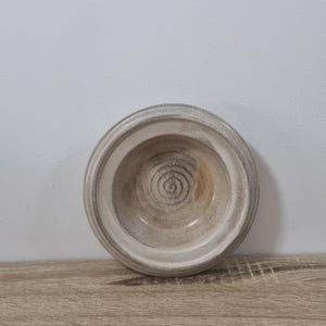 May include: A handmade ceramic bowl with a circular design. The bowl is a neutral colour with a light brown interior and a textured, off-white exterior. The bowl sits on a wooden surface.