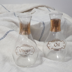 May include: Two clear glass bottles with gold accents and labels. The bottle on the left says "Vinegar" and the bottle on the right says "Oil".