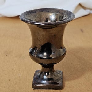 May include: A small, metallic-looking vase with a flared rim and a square base. The vase has a dark, reflective surface with a bronze-like appearance. The shape is reminiscent of a chalice or small urn.