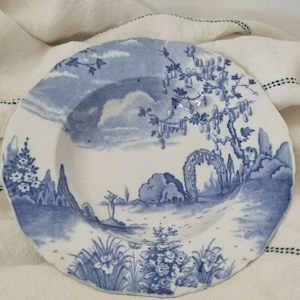 May include: A blue and white ceramic plate with a detailed landscape design. The plate features a garden scene with trees, clouds, and architectural elements. The plate has a scalloped edge. The plate is set against a white cloth.