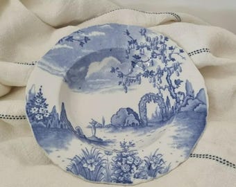 May include: A white and blue ceramic plate with a landscape design. The plate features a detailed blue illustration of a garden scene with trees, clouds, and architectural elements. The plate has a scalloped edge and is set against a white cloth.