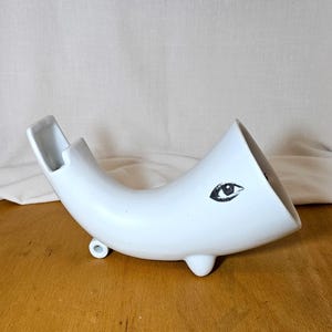 May include: A white ceramic sculpture in a horn shape, with a black eye motif. The sculpture has a small, rounded foot and a rectangular opening at the top. It is placed on a wooden surface. The sculpture measures approximately 20 cm in length.