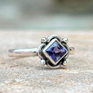 May include: A silver ring with a square, deep purple gemstone at the center. The gemstone is set in a decorative, ornate setting with small, round accents. The ring band is thin and polished.