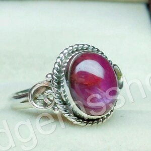 Puede incluir: Un anillo de plata con una piedra preciosa ovalada de color rubí. La piedra está engastada en un bisel decorativo en forma de cuerda. El anillo se presenta en una caja de joyería roja.