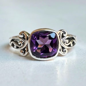 Op de afbeelding: Een zilveren ring met een vierkante, dieppaarse edelsteen. De ring heeft sierlijke, wervelende ontwerpen aan weerszijden van de edelsteen. De ring is geplaatst tegen een witte achtergrond.